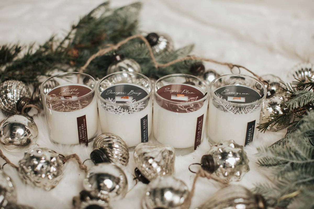 Four white scented candles in clear glass jars arranged on a white fabric surface with silver ornaments and evergreen branches around them. The candle labels show the brand 'Bourgeon Bliss' and scent names including 'Winter Breeze', 'Snowfall', and 'Winter Dreams'.
