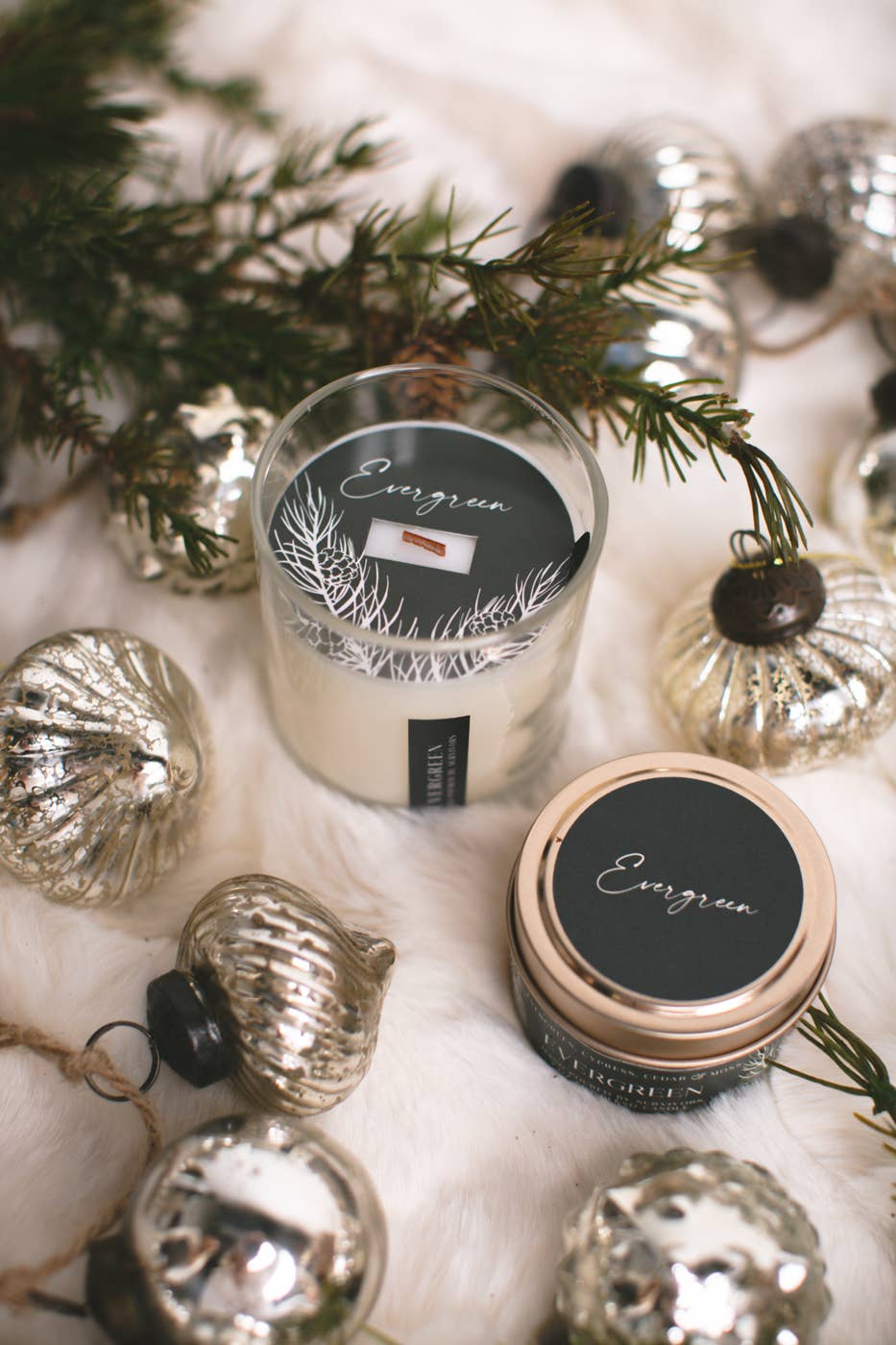 Two candles from the brand Evergreen, one in a clear glass jar and one in a tin with a rose gold lid, both featuring black labels with white pine tree designs. They are styled on a white faux fur surface with silver ornaments and pine branches.